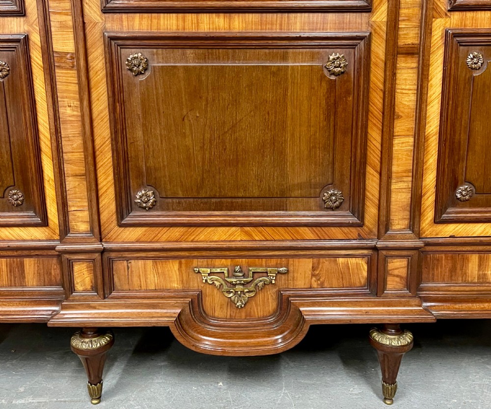 An antique French three-door bookcase crafted from kingwood and mahogany, featuring wire-mesh upper doors, lower timber panel doors, turned legs, and carved decorative crests. View IX