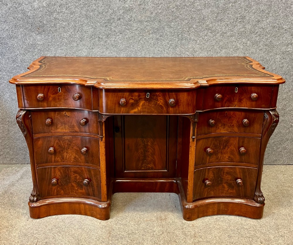 A Victorian figured mahogany pedestal desk featuring a shaped gilded leather surface, serpentine drawer fronts, and turned wooden knobs.