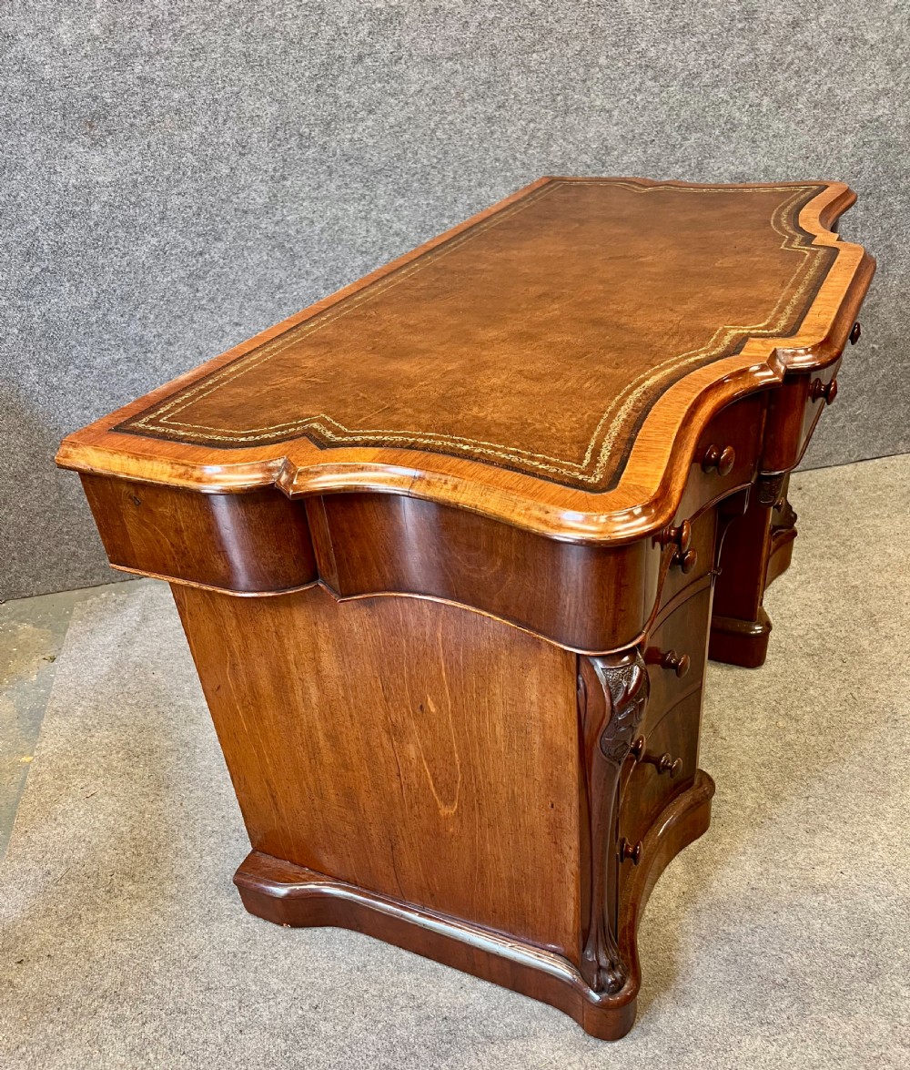A Victorian figured mahogany pedestal desk featuring a shaped gilded leather surface, serpentine drawer fronts, and turned wooden knobs. View V