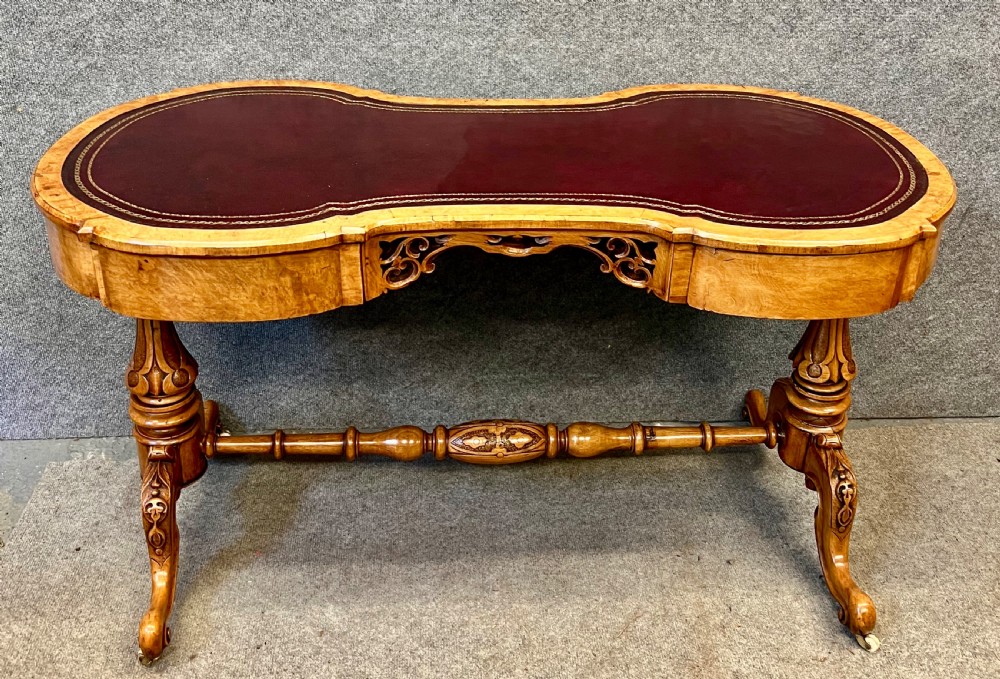 A Victorian burr walnut kidney-shaped writing desk featuring a burgundy leather writing surface, openwork carved frieze, turned central stretcher, and cabriole legs on brass castors.