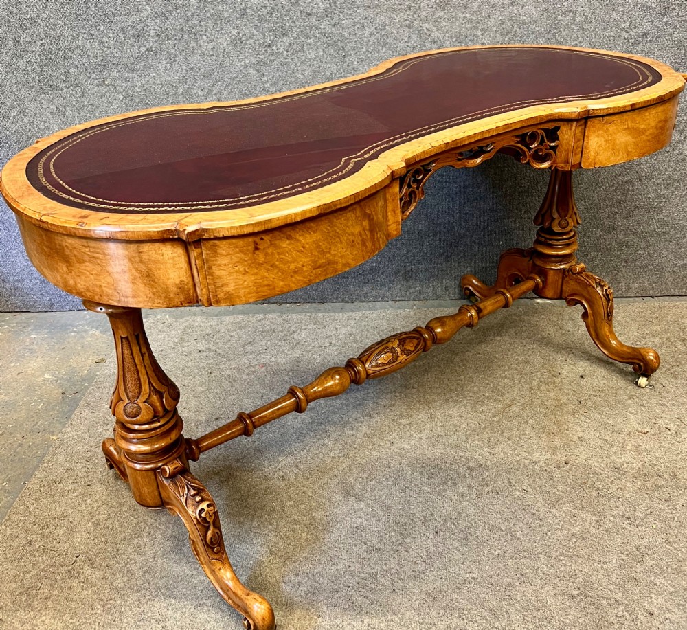 A Victorian burr walnut kidney-shaped writing desk featuring a burgundy leather writing surface, openwork carved frieze, turned central stretcher, and cabriole legs on brass castors. View II