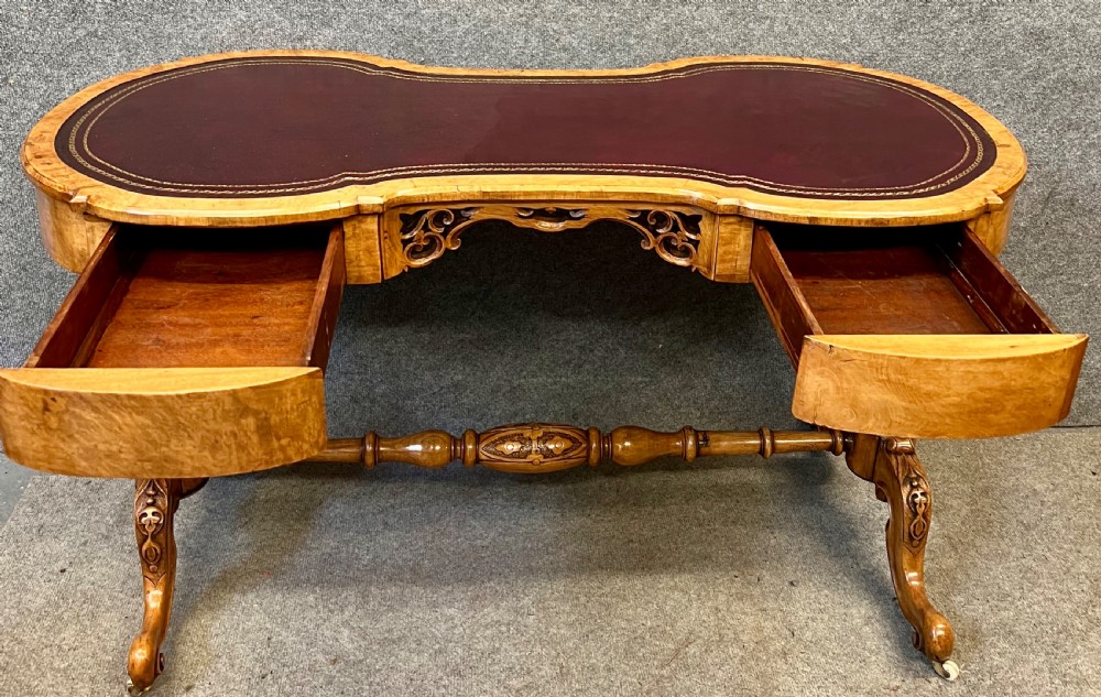 A Victorian burr walnut kidney-shaped writing desk featuring a burgundy leather writing surface, openwork carved frieze, turned central stretcher, and cabriole legs on brass castors. View III