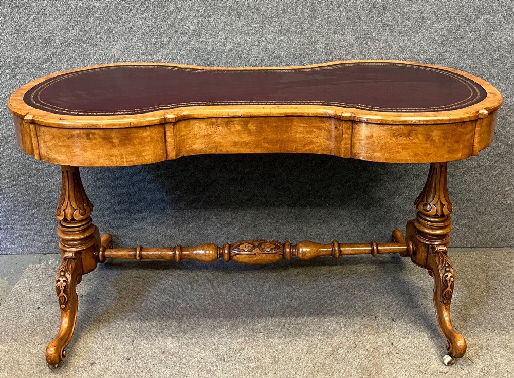 A Victorian burr walnut kidney-shaped writing desk featuring a burgundy leather writing surface, openwork carved frieze, turned central stretcher, and cabriole legs on brass castors. View V