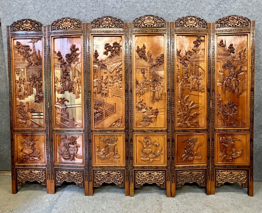 A six-panel wooden folding screen featuring high-relief carved scenes of figures, buildings, and plants framed by dark fretwork borders.
