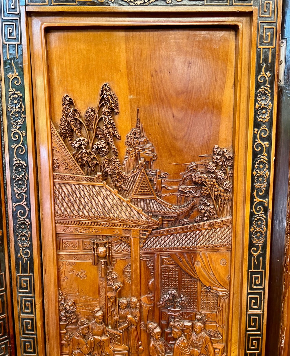A six-panel wooden folding screen featuring high-relief carved scenes of figures, buildings, and plants framed by dark fretwork borders. View VI