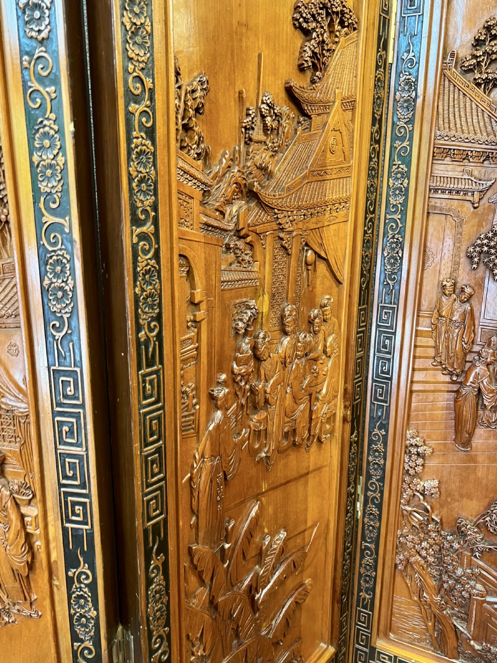 A six-panel wooden folding screen featuring high-relief carved scenes of figures, buildings, and plants framed by dark fretwork borders. View VII