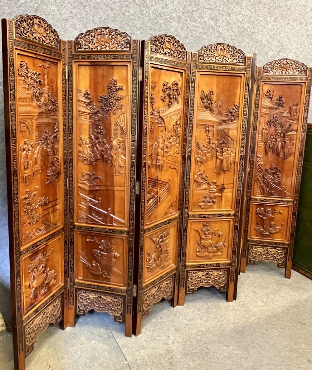 A six-panel wooden folding screen featuring high-relief carved scenes of figures, buildings, and plants framed by dark fretwork borders. View XII