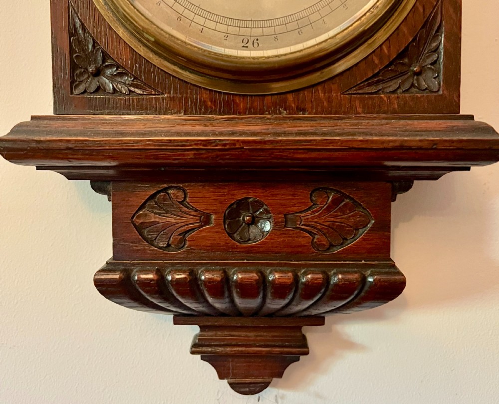 A Victorian carved dark oak wall barometer featuring an upper mercury thermometer flanked by fluted columns and a lower circular brass-rimmed aneroid dial. View V