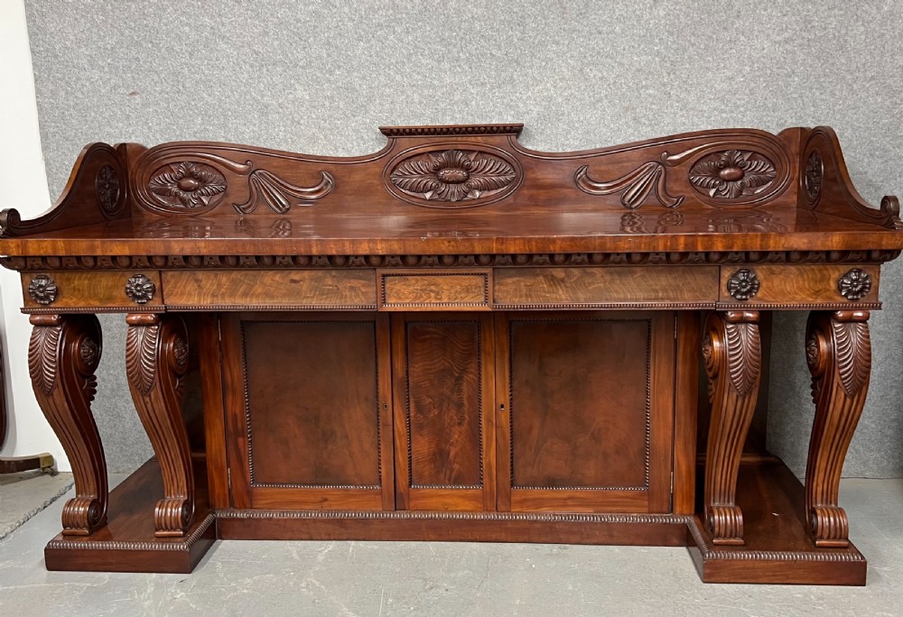 A large Regency mahogany sideboard featuring carved floral scrolls on the raised backboard, heavy carved scroll supports, and panelled cupboard doors.
