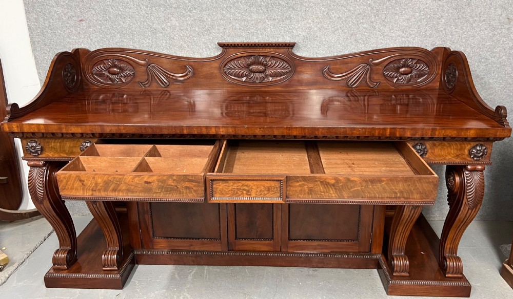 A large Regency mahogany sideboard featuring carved floral scrolls on the raised backboard, heavy carved scroll supports, and panelled cupboard doors. View II