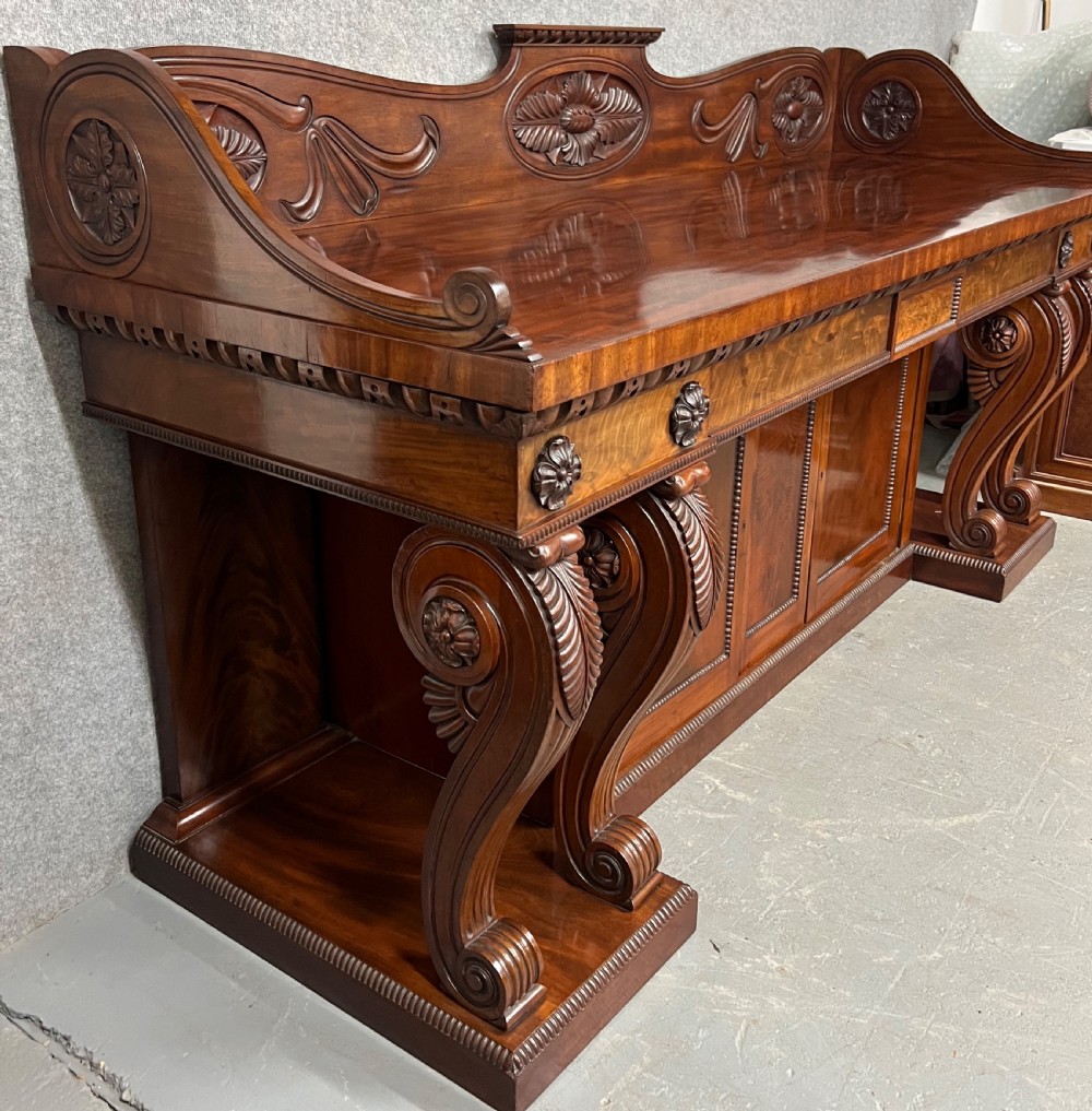 A large Regency mahogany sideboard featuring carved floral scrolls on the raised backboard, heavy carved scroll supports, and panelled cupboard doors. View VIII