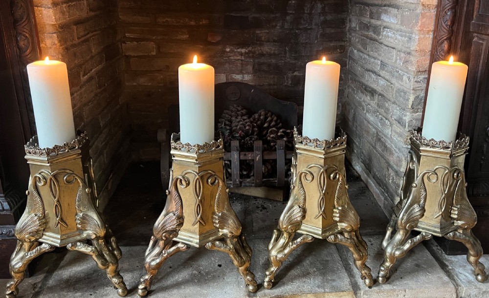 A set of four large ornate cast brass candlesticks featuring pierced crenellated rims, square pedestal bases, and sculpted winged legs ending in hoof feet.