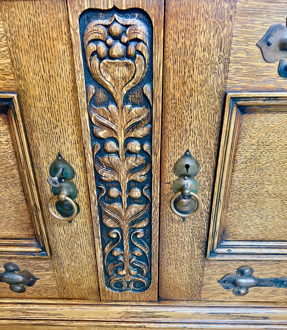 A golden oak Arts and Crafts sideboard featuring an upper shelf, a central repousse copper relief panel above two mirrors, and a lower cabinet with iron strap hinges and carved details. View IX