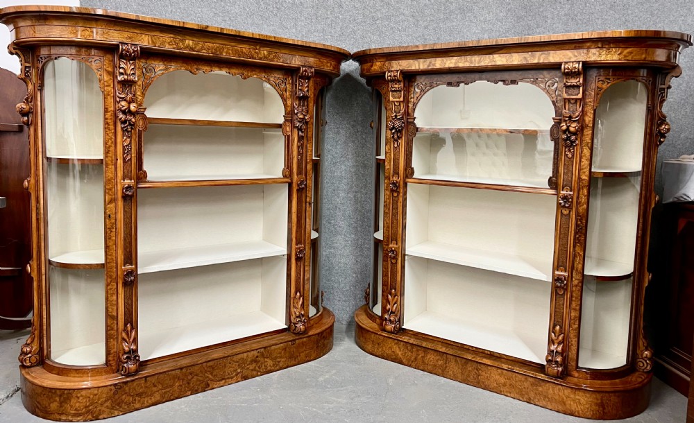 A pair of matching Victorian burr walnut cabinets featuring carved foliate detailing, open central shelving, and curved glass side compartments. View XIII