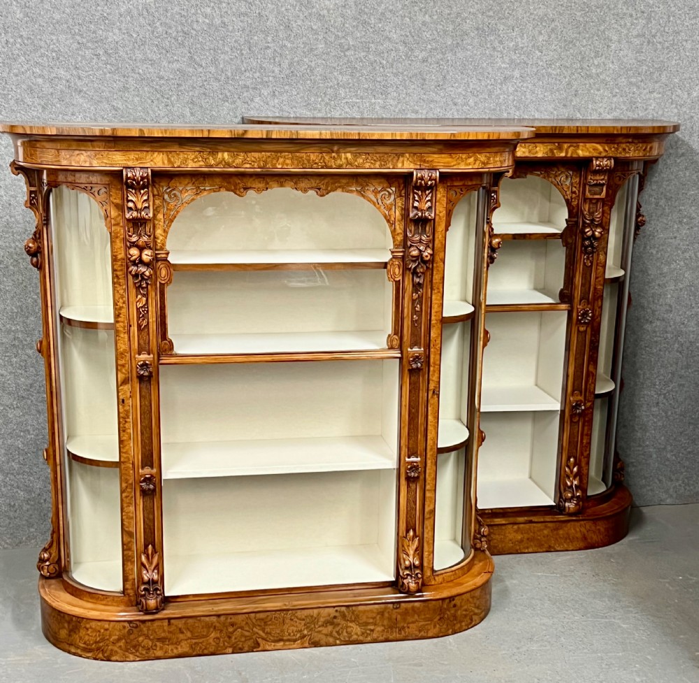 A pair of matching Victorian burr walnut cabinets featuring carved foliate detailing, open central shelving, and curved glass side compartments. View II