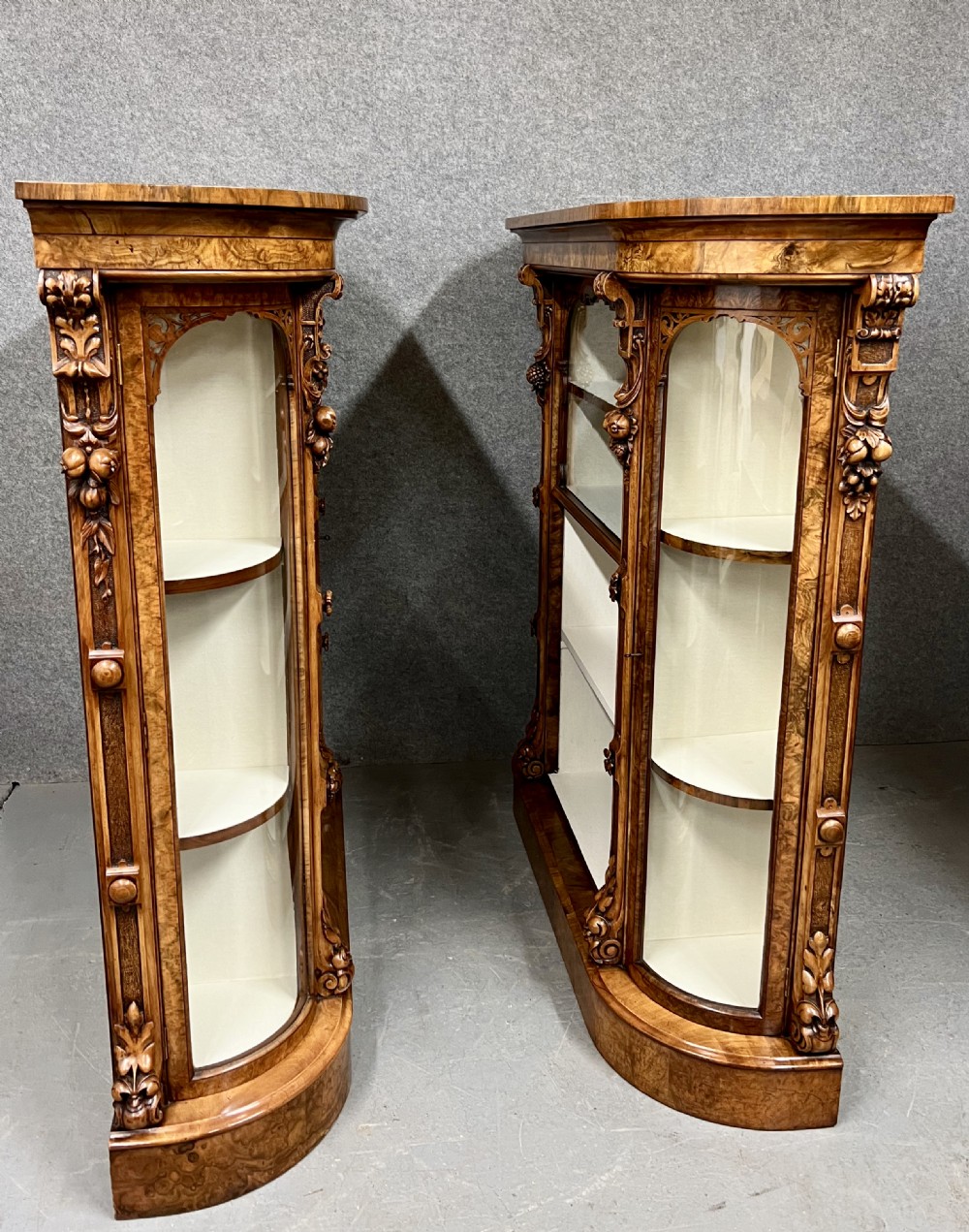 A pair of matching Victorian burr walnut cabinets featuring carved foliate detailing, open central shelving, and curved glass side compartments. View V