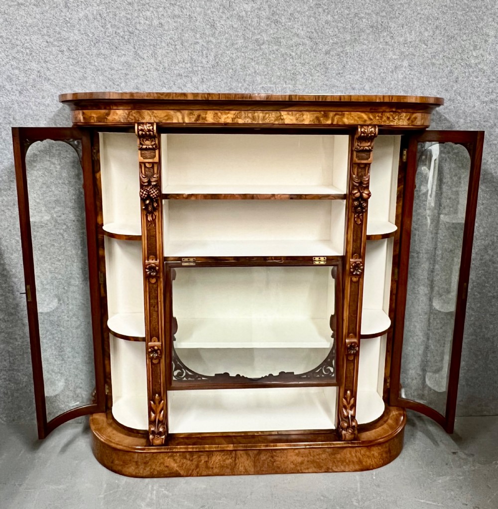 A pair of matching Victorian burr walnut cabinets featuring carved foliate detailing, open central shelving, and curved glass side compartments. View VI