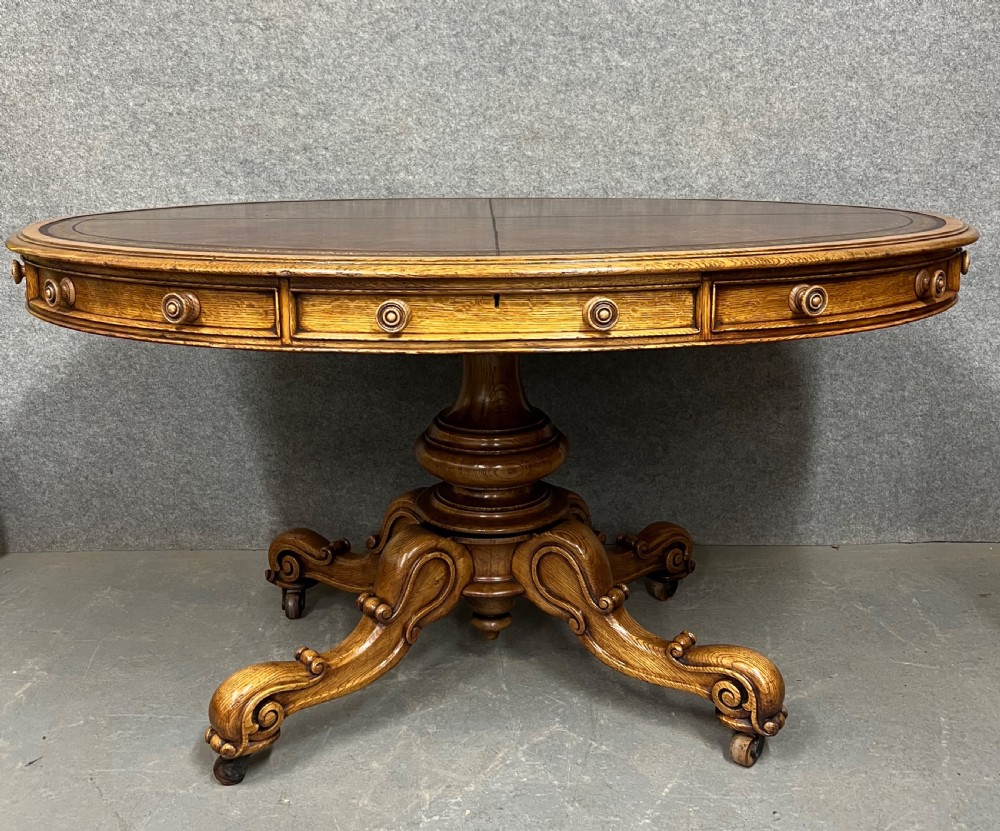 An antique Victorian oval oak library table featuring a leather inset top, perimeter frieze drawers, and a heavy carved pedestal base with scrolled legs on castors.