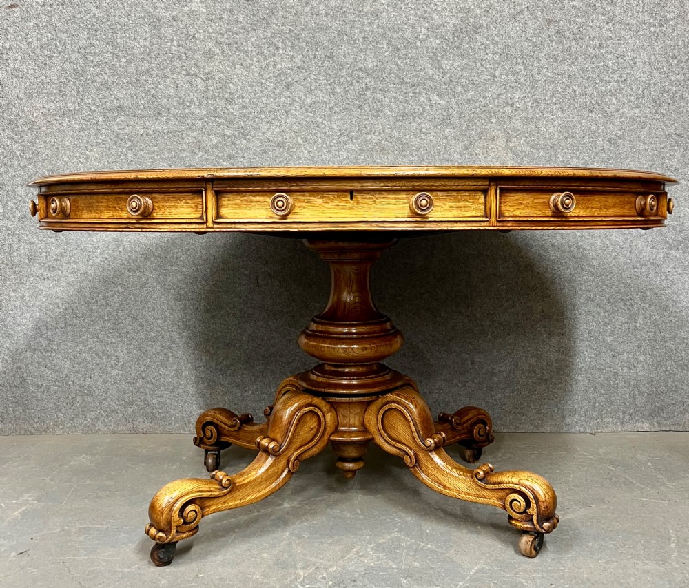 An antique Victorian oval oak library table featuring a leather inset top, perimeter frieze drawers, and a heavy carved pedestal base with scrolled legs on castors. View II