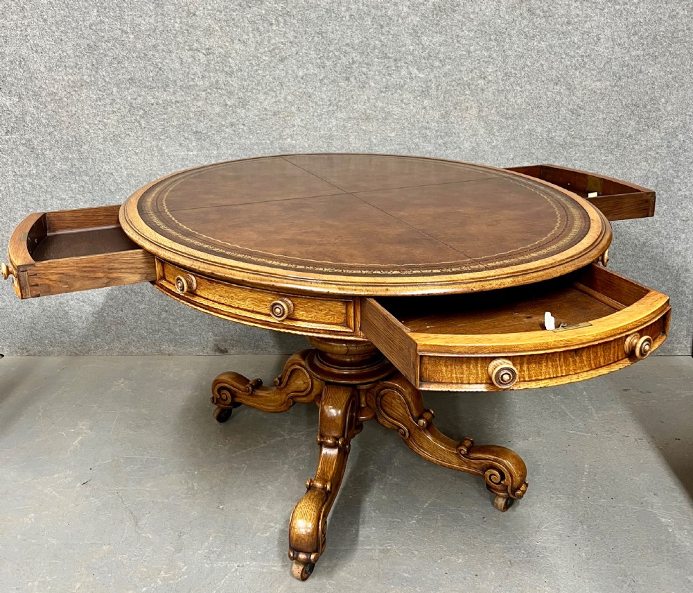 An antique Victorian oval oak library table featuring a leather inset top, perimeter frieze drawers, and a heavy carved pedestal base with scrolled legs on castors. View III