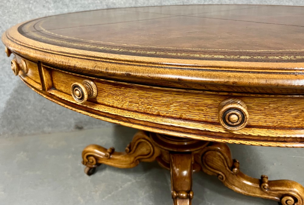 An antique Victorian oval oak library table featuring a leather inset top, perimeter frieze drawers, and a heavy carved pedestal base with scrolled legs on castors. View V