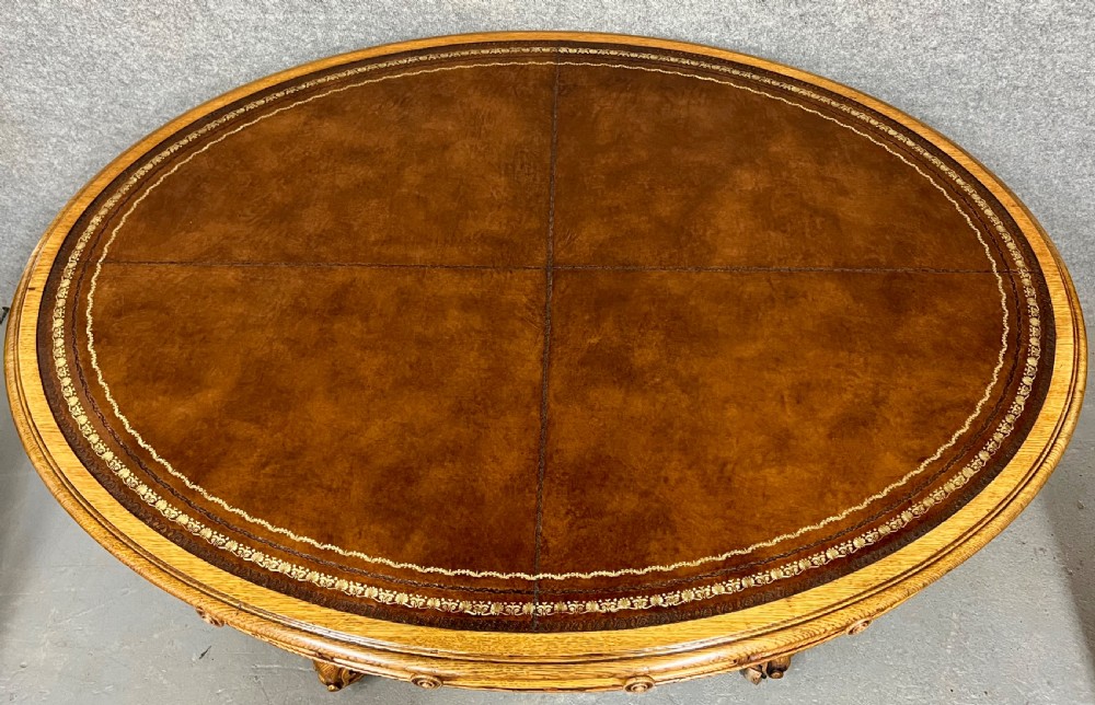 An antique Victorian oval oak library table featuring a leather inset top, perimeter frieze drawers, and a heavy carved pedestal base with scrolled legs on castors. View VI
