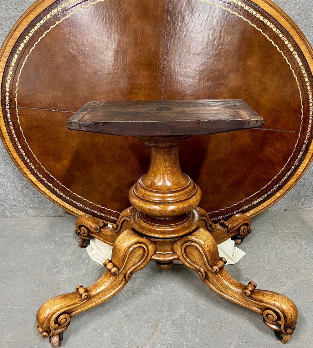 An antique Victorian oval oak library table featuring a leather inset top, perimeter frieze drawers, and a heavy carved pedestal base with scrolled legs on castors. View VIII