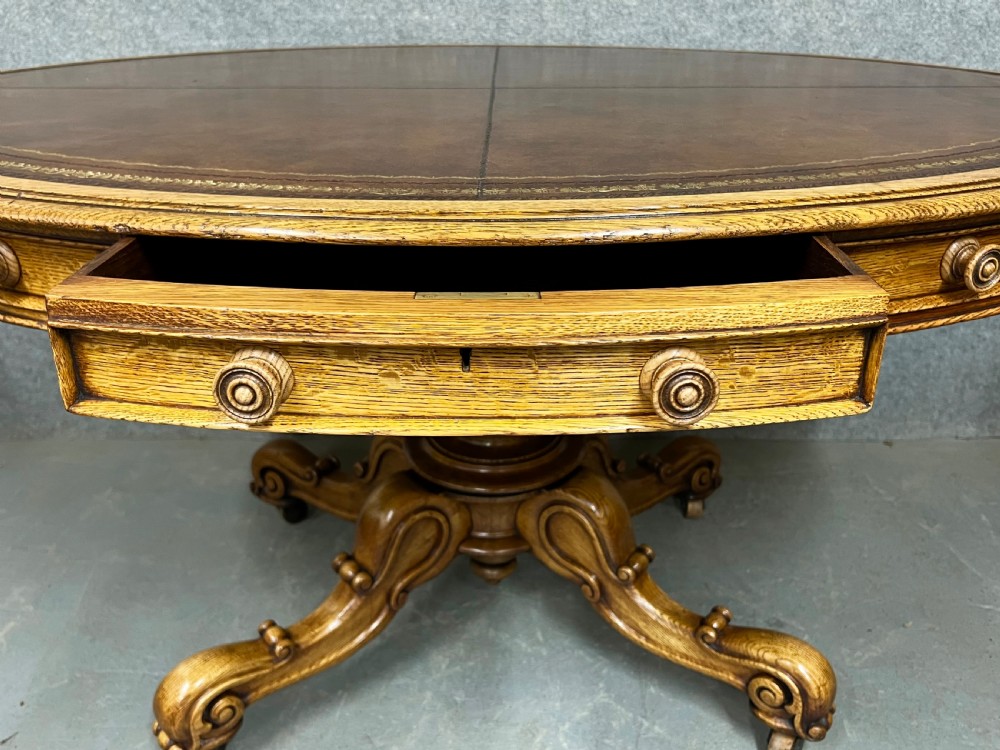 An antique Victorian oval oak library table featuring a leather inset top, perimeter frieze drawers, and a heavy carved pedestal base with scrolled legs on castors. View IX