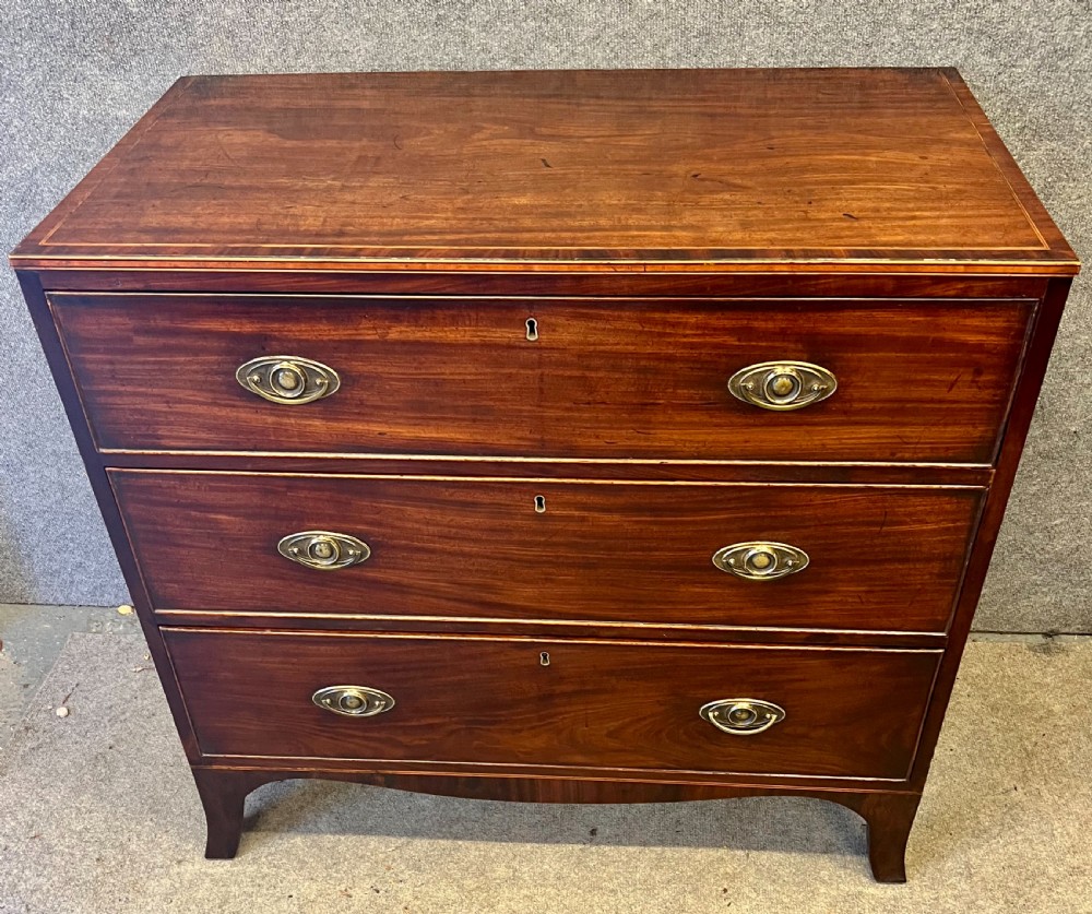 A George III mahogany chest of drawers featuring three graduated drawers with brass oval handles and line inlay on shaped bracket feet.