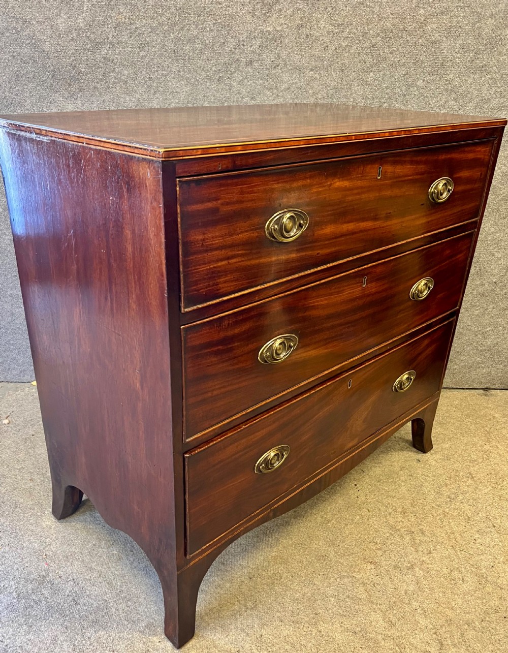 A George III mahogany chest of drawers featuring three graduated drawers with brass oval handles and line inlay on shaped bracket feet. View II