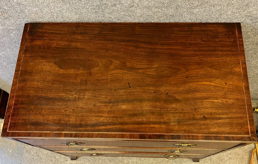 A George III mahogany chest of drawers featuring three graduated drawers with brass oval handles and line inlay on shaped bracket feet. View V