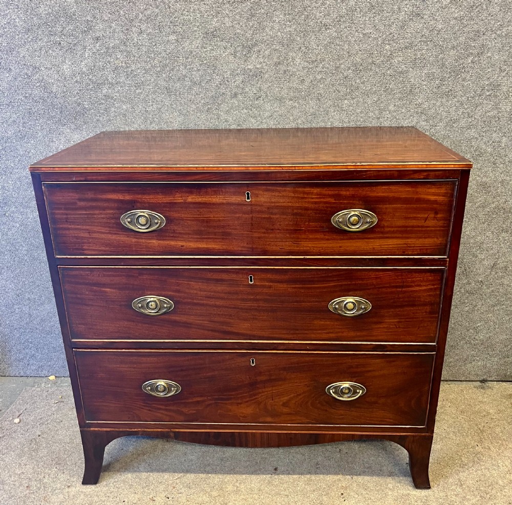 A George III mahogany chest of drawers featuring three graduated drawers with brass oval handles and line inlay on shaped bracket feet. View IX