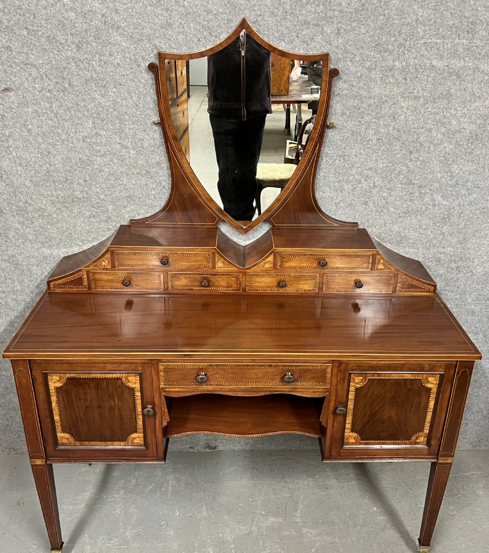 An antique Sheraton Revival mahogany dressing table featuring a central shield-shaped mirror, satinwood inlay, multiple small drawers, and square tapered legs on castors. View II