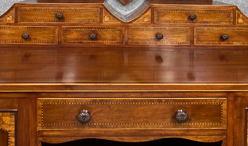An antique Sheraton Revival mahogany dressing table featuring a central shield-shaped mirror, satinwood inlay, multiple small drawers, and square tapered legs on castors. View III