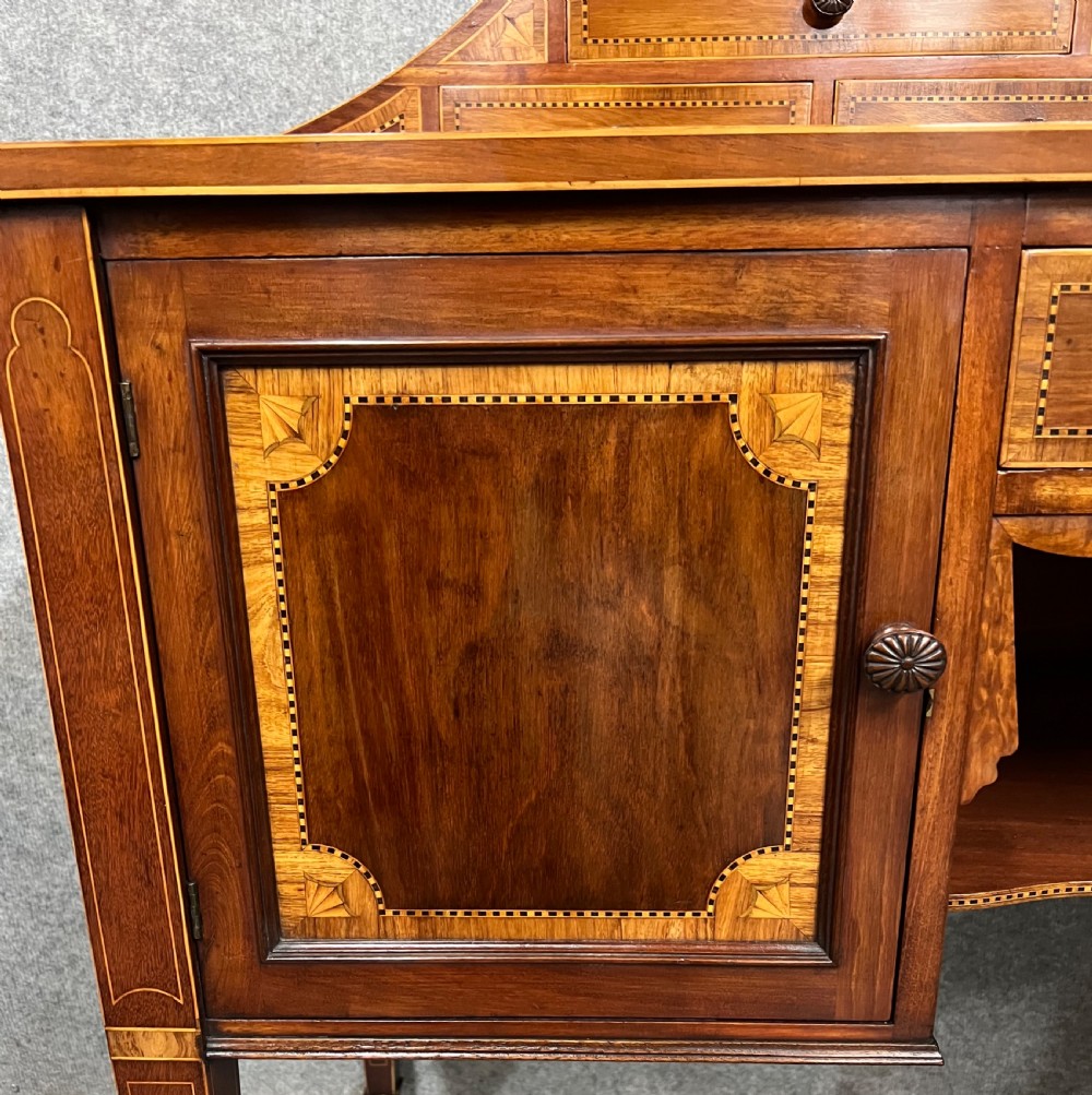 An antique Sheraton Revival mahogany dressing table featuring a central shield-shaped mirror, satinwood inlay, multiple small drawers, and square tapered legs on castors. View IV