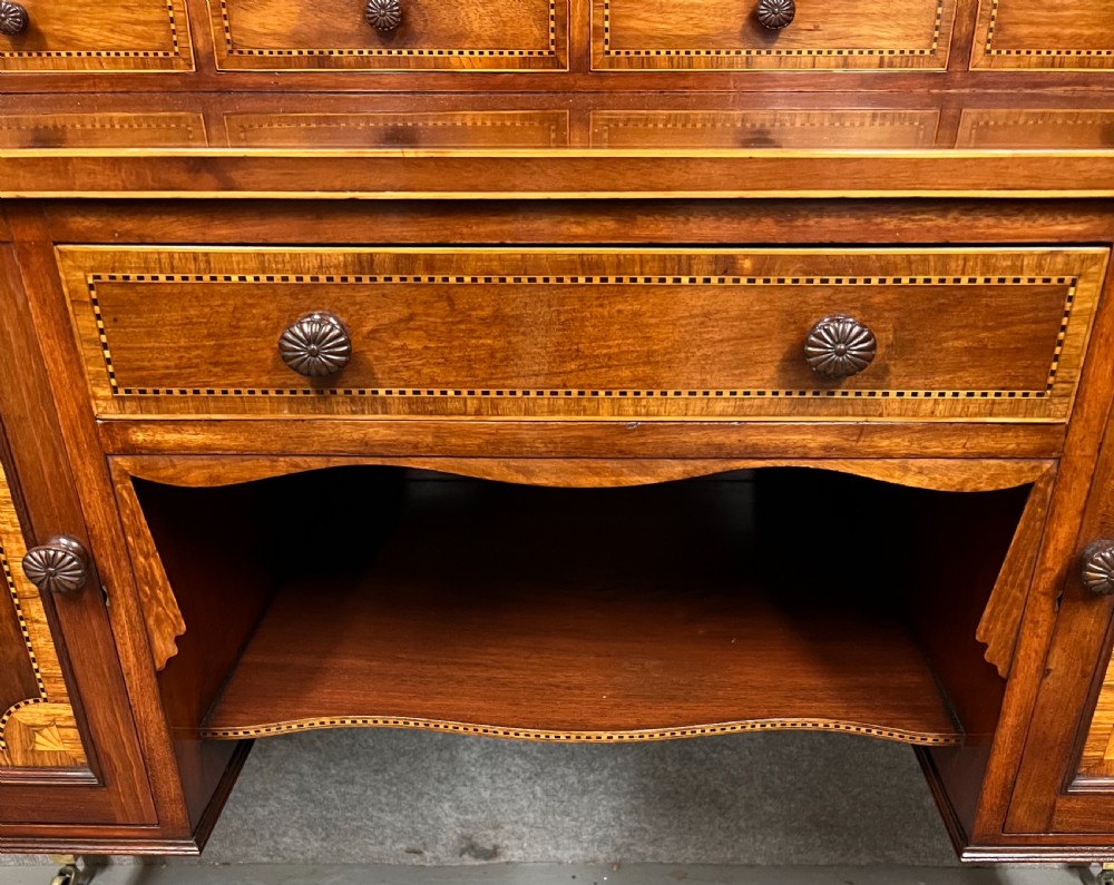 An antique Sheraton Revival mahogany dressing table featuring a central shield-shaped mirror, satinwood inlay, multiple small drawers, and square tapered legs on castors. View V