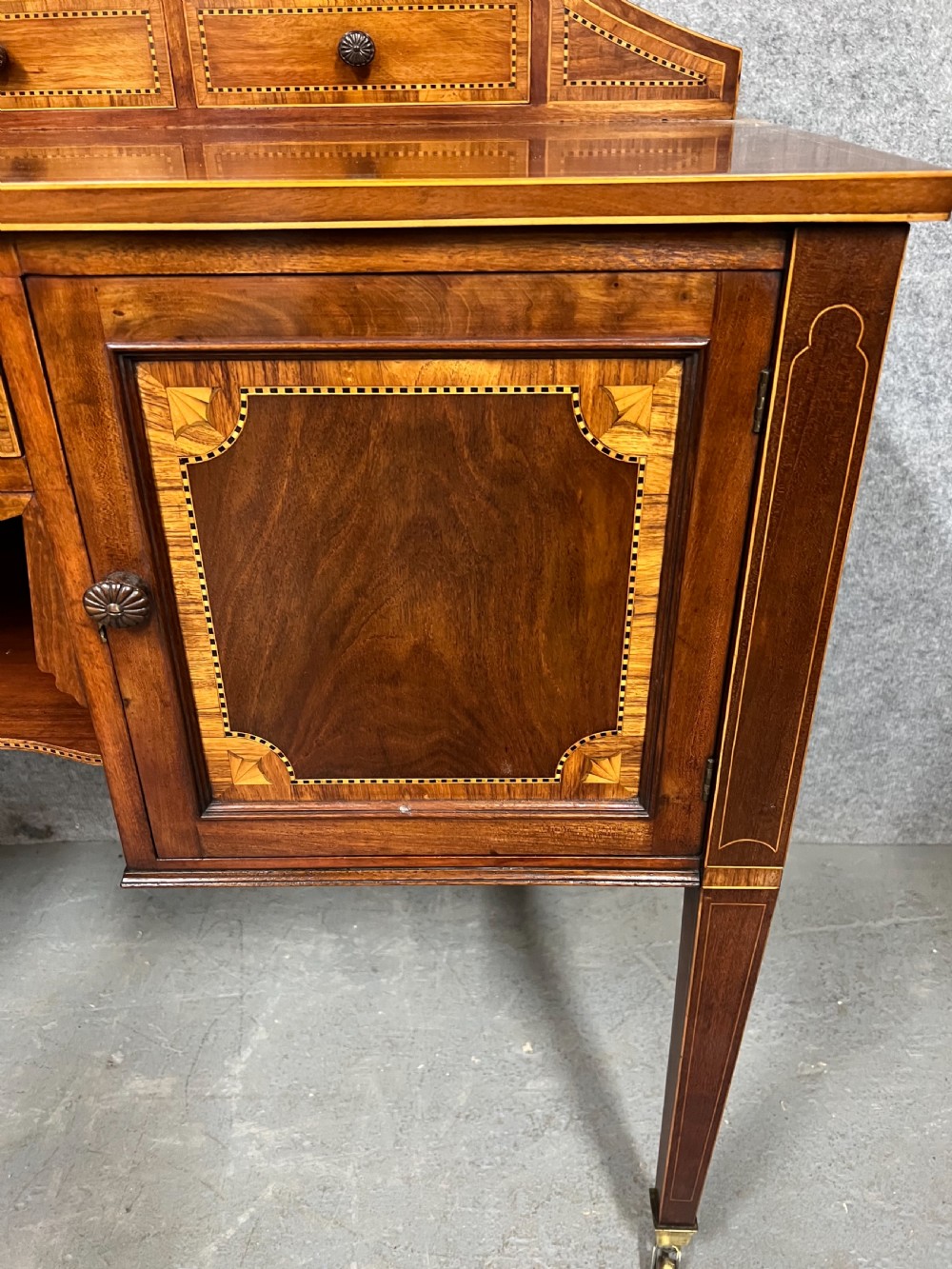 An antique Sheraton Revival mahogany dressing table featuring a central shield-shaped mirror, satinwood inlay, multiple small drawers, and square tapered legs on castors. View VI