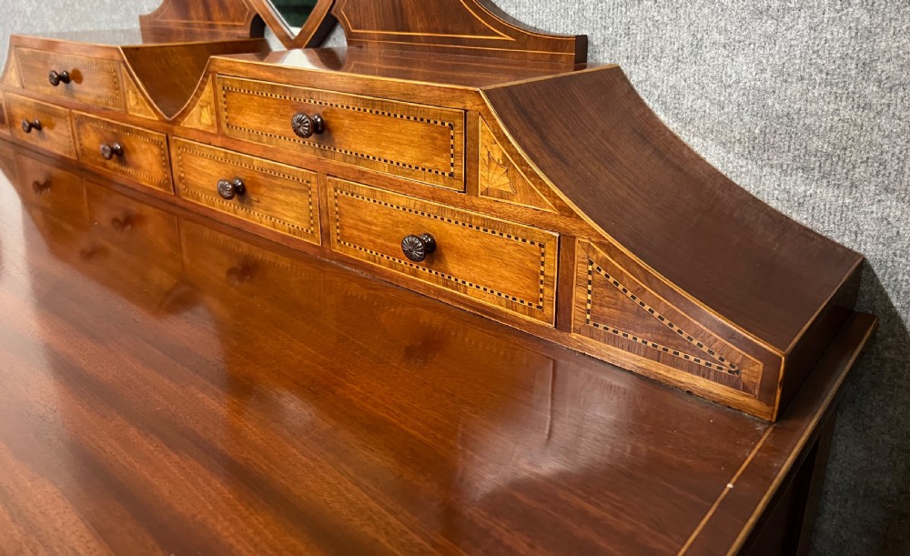 An antique Sheraton Revival mahogany dressing table featuring a central shield-shaped mirror, satinwood inlay, multiple small drawers, and square tapered legs on castors. View VII