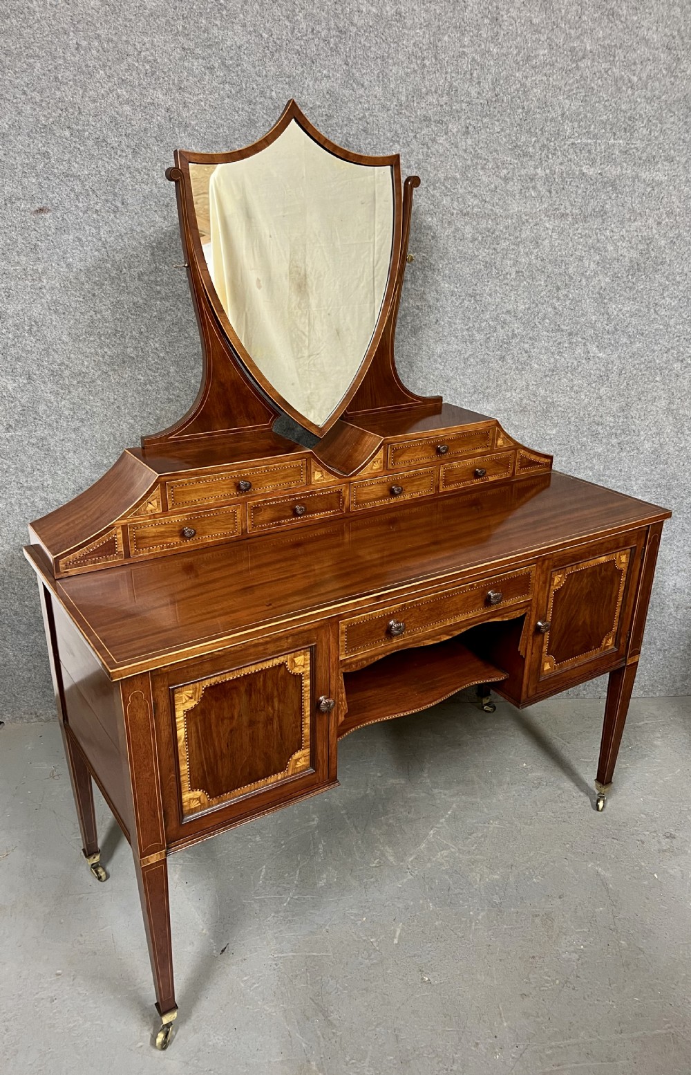 An antique Sheraton Revival mahogany dressing table featuring a central shield-shaped mirror, satinwood inlay, multiple small drawers, and square tapered legs on castors. View IX