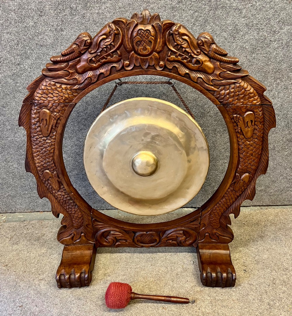 A hand-carved dark wood circular gong stand featuring twin dragon motifs and claw feet, suspending a central brass gong with a red textile mallet below.