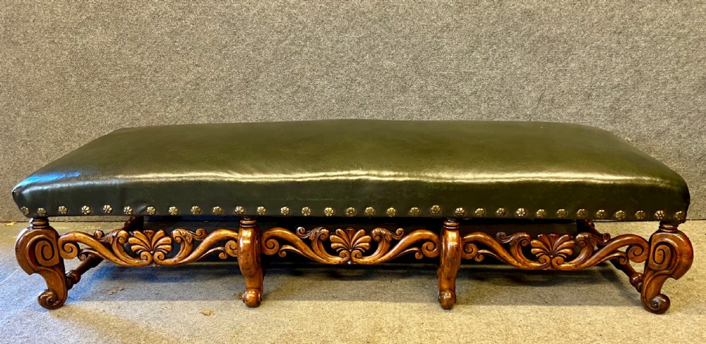 A long, low carved walnut stool featuring dark green leather upholstery secured with decorative brass studs along the apron.