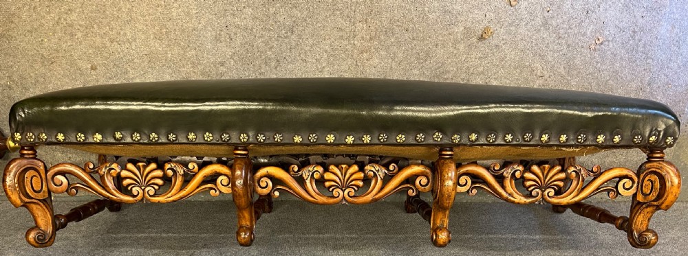 A long, low carved walnut stool featuring dark green leather upholstery secured with decorative brass studs along the apron. View II