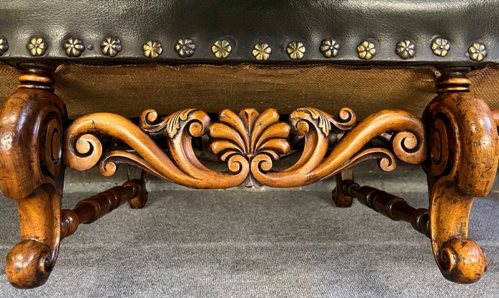 A long, low carved walnut stool featuring dark green leather upholstery secured with decorative brass studs along the apron. View III
