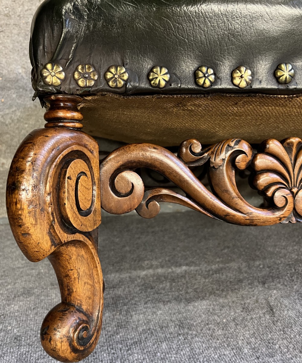 A long, low carved walnut stool featuring dark green leather upholstery secured with decorative brass studs along the apron. View IV