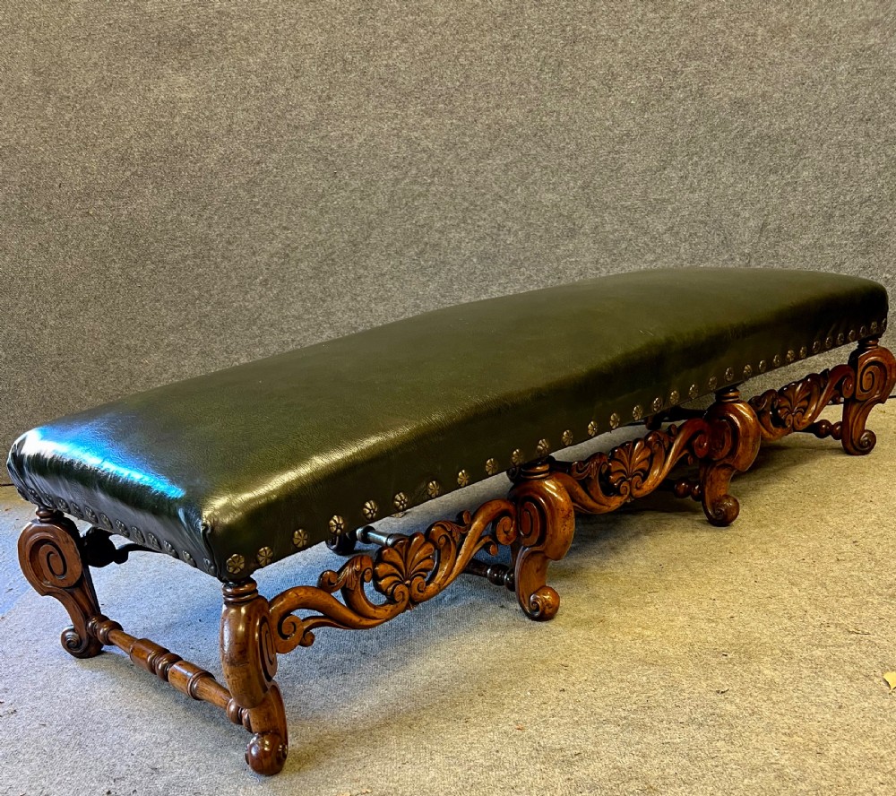 A long, low carved walnut stool featuring dark green leather upholstery secured with decorative brass studs along the apron. View V
