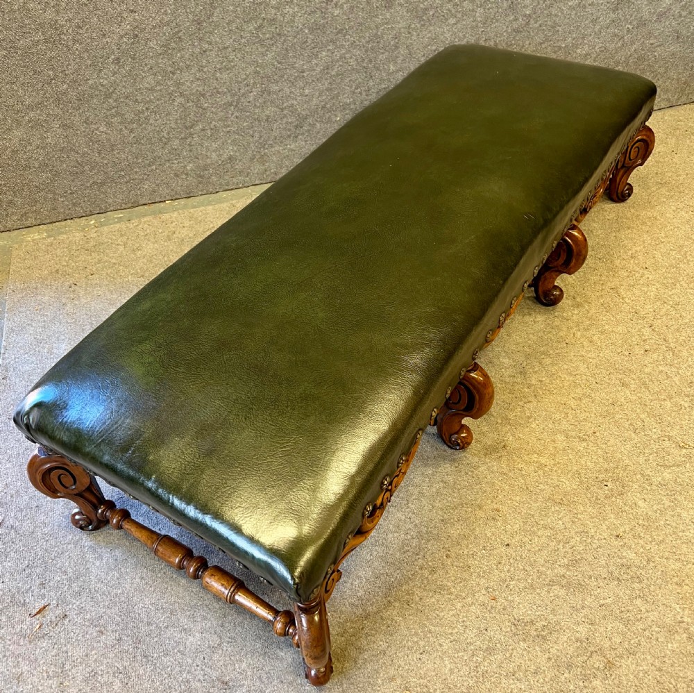 A long, low carved walnut stool featuring dark green leather upholstery secured with decorative brass studs along the apron. View VI