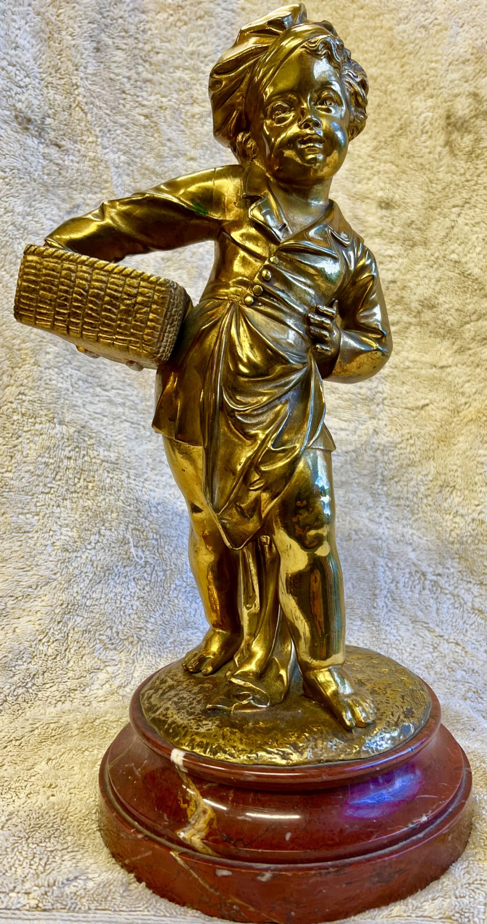 A gilt bronze sculpture depicting a young girl holding a woven basket on a circular red marble base.