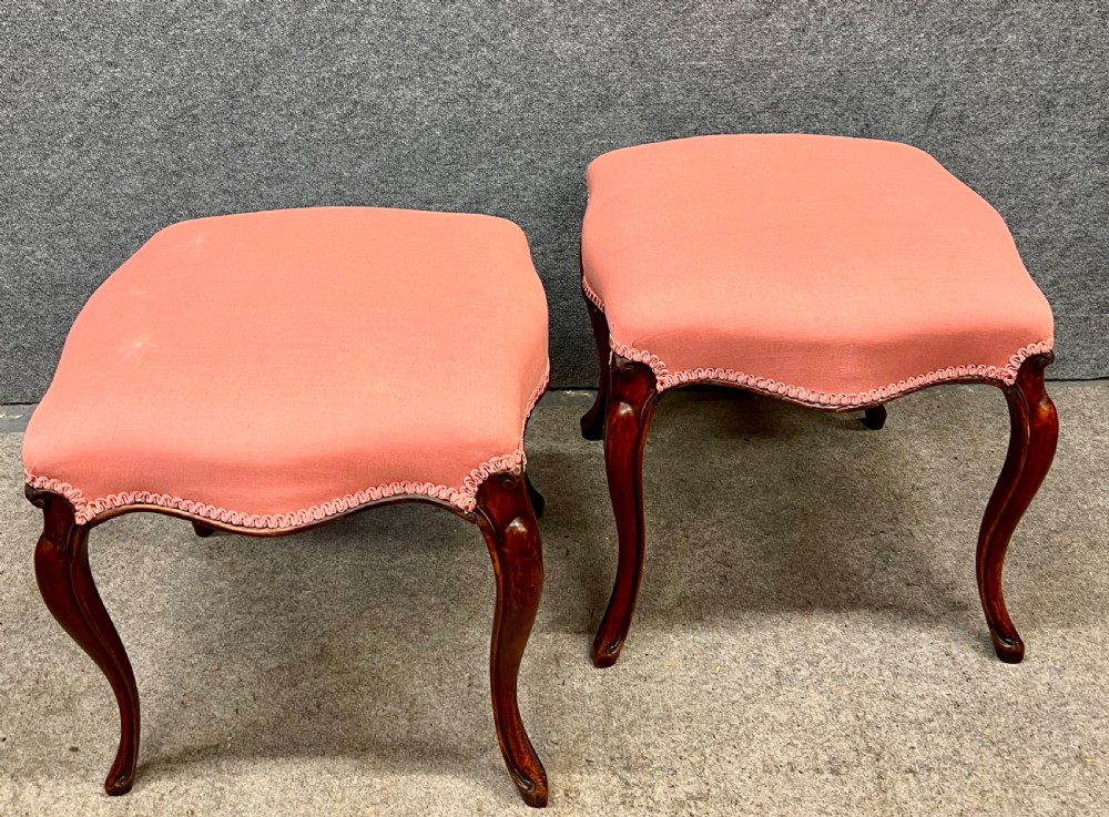 A pair of Victorian footstools with pink upholstered seats, decorative braided gimp trim, and carved dark wood cabriole legs.
