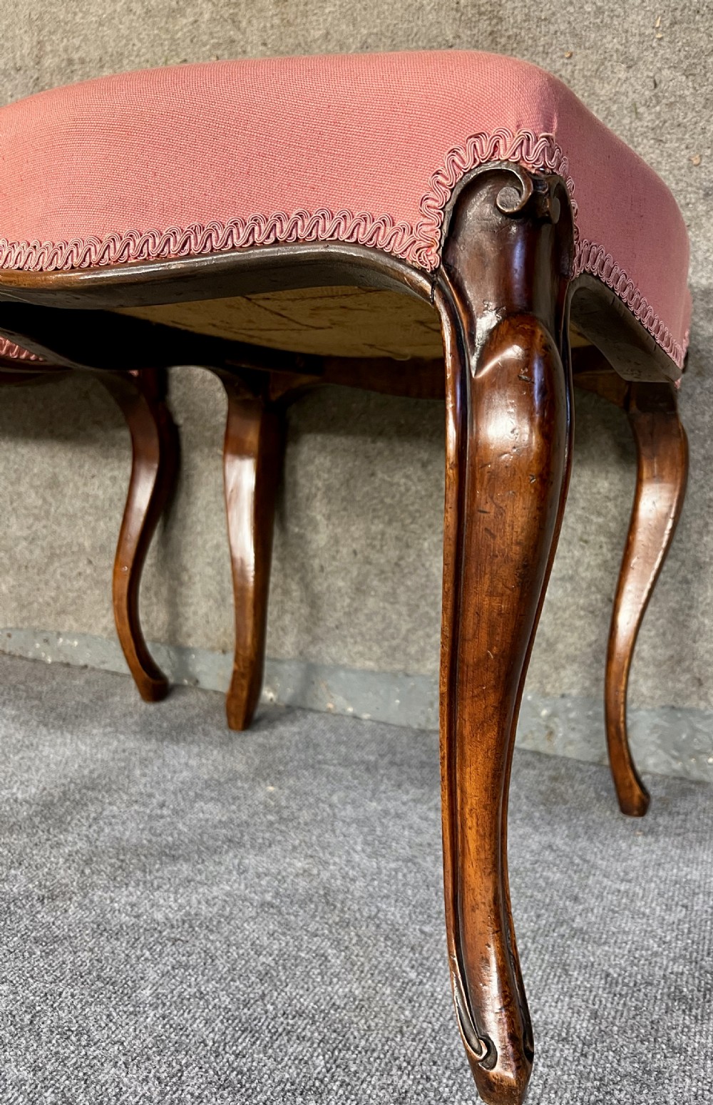 A pair of Victorian footstools with pink upholstered seats, decorative braided gimp trim, and carved dark wood cabriole legs. View II