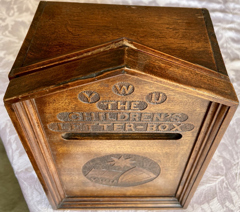 A wall-mounted carved oak letter box with a pointed top, horizontal aperture slot, side metal catch, and carved emblems on the front panel. View II